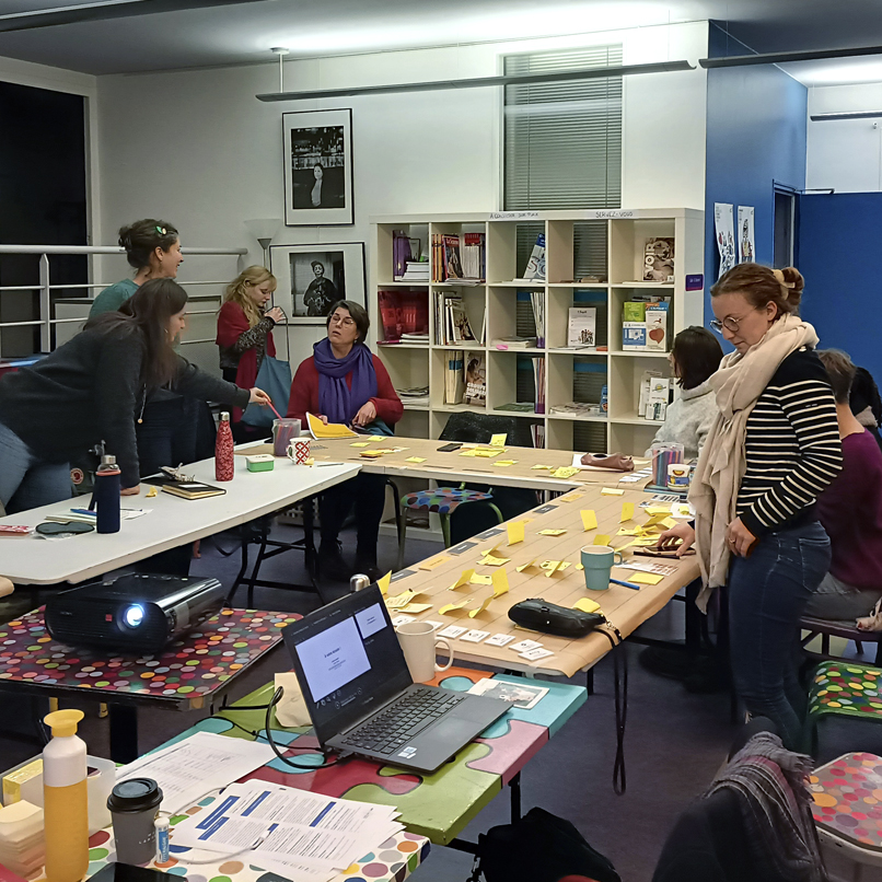 séance de travail, échange entre 6 participantes, dans la salle les tables sont placées en U, il y a des ordinateurs portables, un projecteur, des post it de toutes les couleurs collés sur les tables.