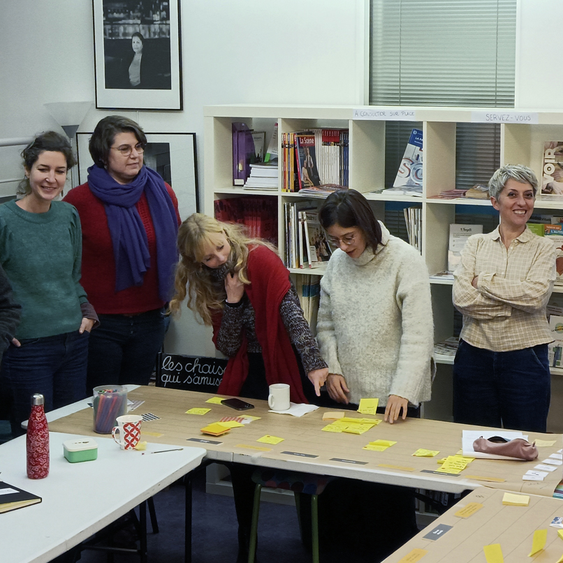 séance de travail, échange entre 5 participantes et Gwenaëlle Brochoire, dans la salle les tables sont placées en U, il y a des ordinateurs portables, un projecteur, des post it de toutes les couleurs collés sur les tables.