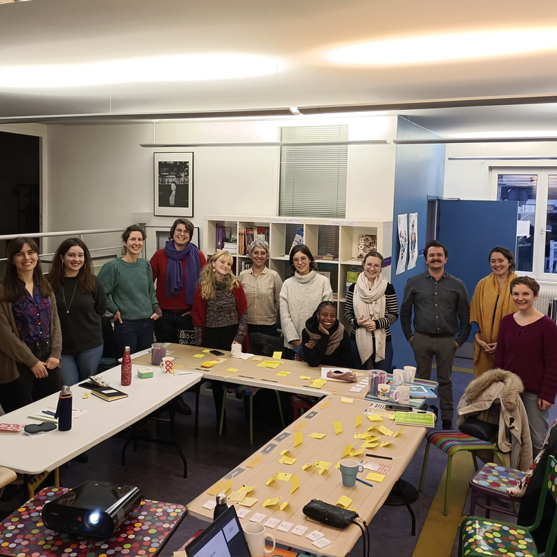 Photo de groupe des 9 participantes et des 3 formateur et formatrices émilie Bougouin, Victor Dobin et Gwenaëlle Brochoire, dans la salle les tables sont placées en U, il y a des ordinateurs portables, un projecteur, des post it de toutes les couleurs collés sur les tables.